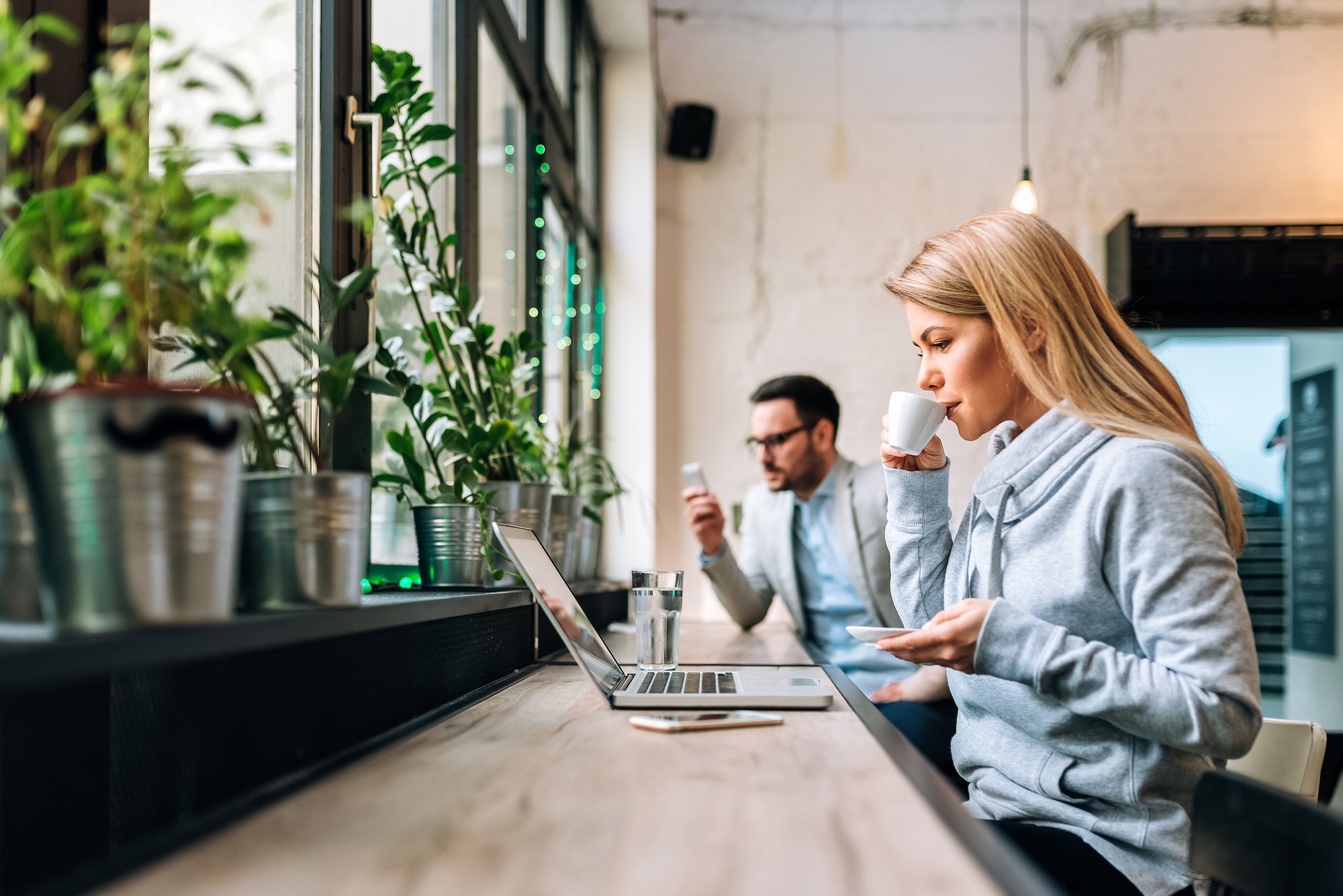 Young woman drinking coffee while working remotely on a laptop with handsome man at the background.