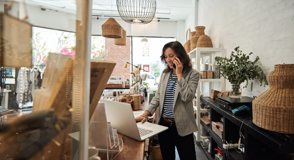 Smiling young Asian woman standing behind a counter in her stylish boutique working on a laptop and talking on a cellphone